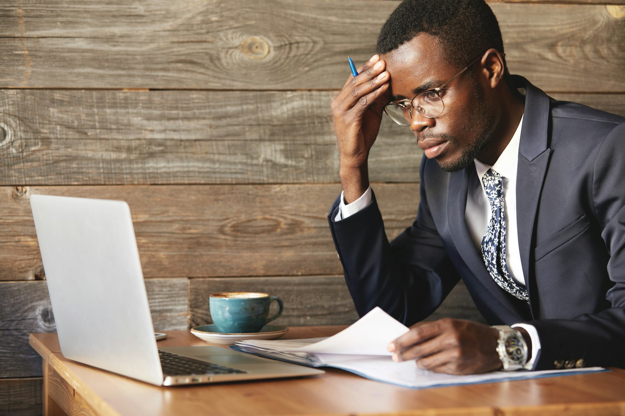 Worried African businessman in official suit checking information in laptop and papers in cosy caf�H